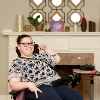 A young brunette woman in a patterned shirt and glasses sits in her wheelchair in front of a fireplace.