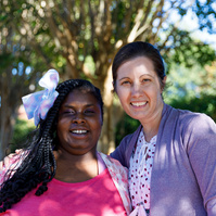 Mother and daughter stand outside under a tree on a sunny day. 