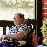 A young brunette woman in a patterned shirt and glasses sits on a screened porch in her wheelchair. 