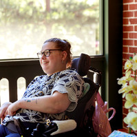 A young brunette woman in a patterned shirt and glasses sits on a screened porch in her wheelchair. 