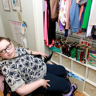 A young brunette woman in a patterned shirt and glasses sits in front of her closet full of colorful clothes.