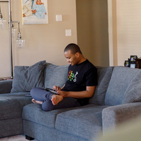 A young African American man sits on a blue sofa and looks at a cell phone