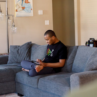 A young African American man sits on a blue sofa and looks at a cell phone