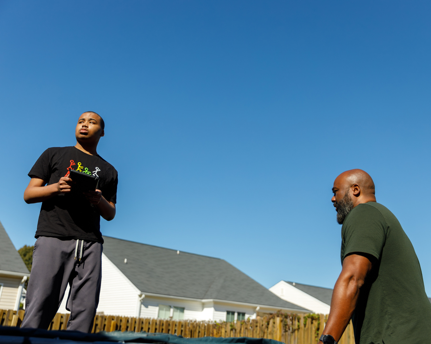 A young man stands on a trampoline in front of a blue sky while his father stands near him. 