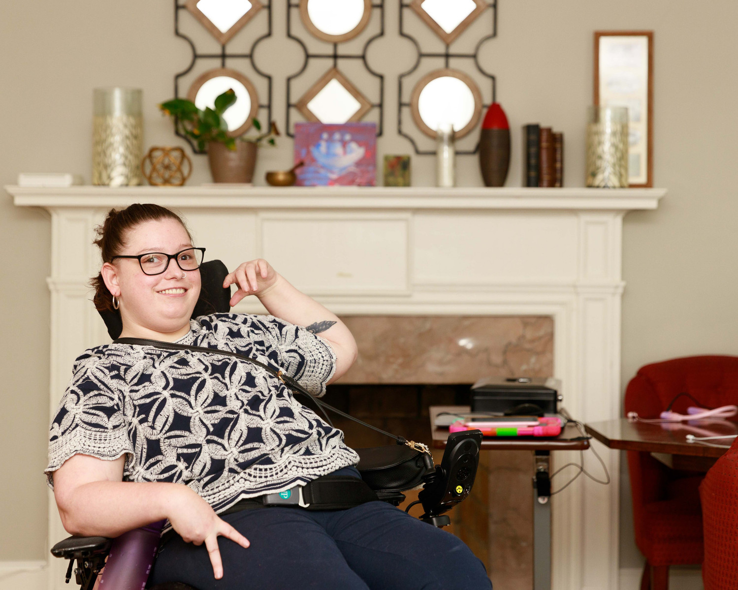 A young brunette woman in a patterned shirt and glasses sits in her wheelchair in front of a fireplace.