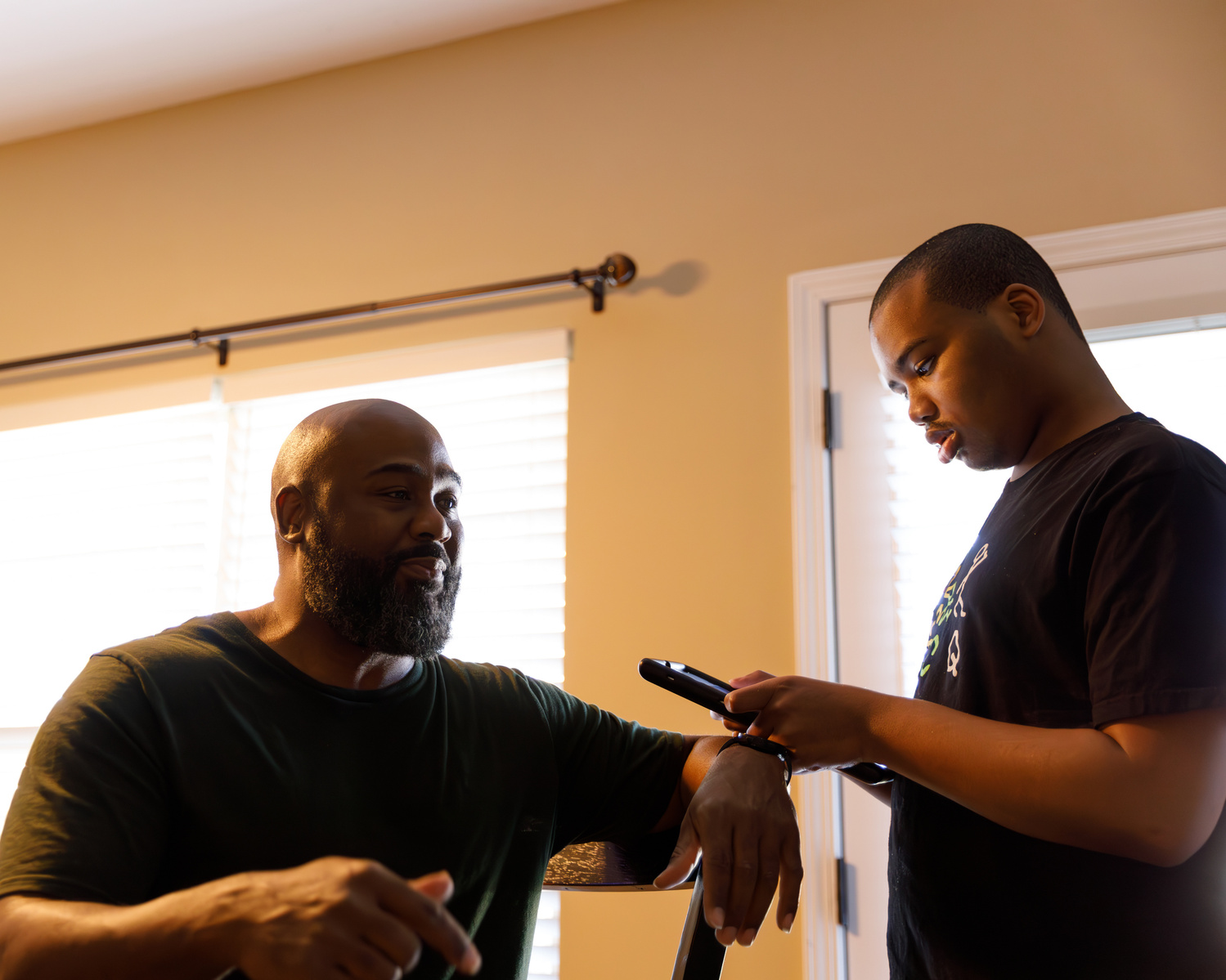 A father and son talk as the son looks at a cell phone. 