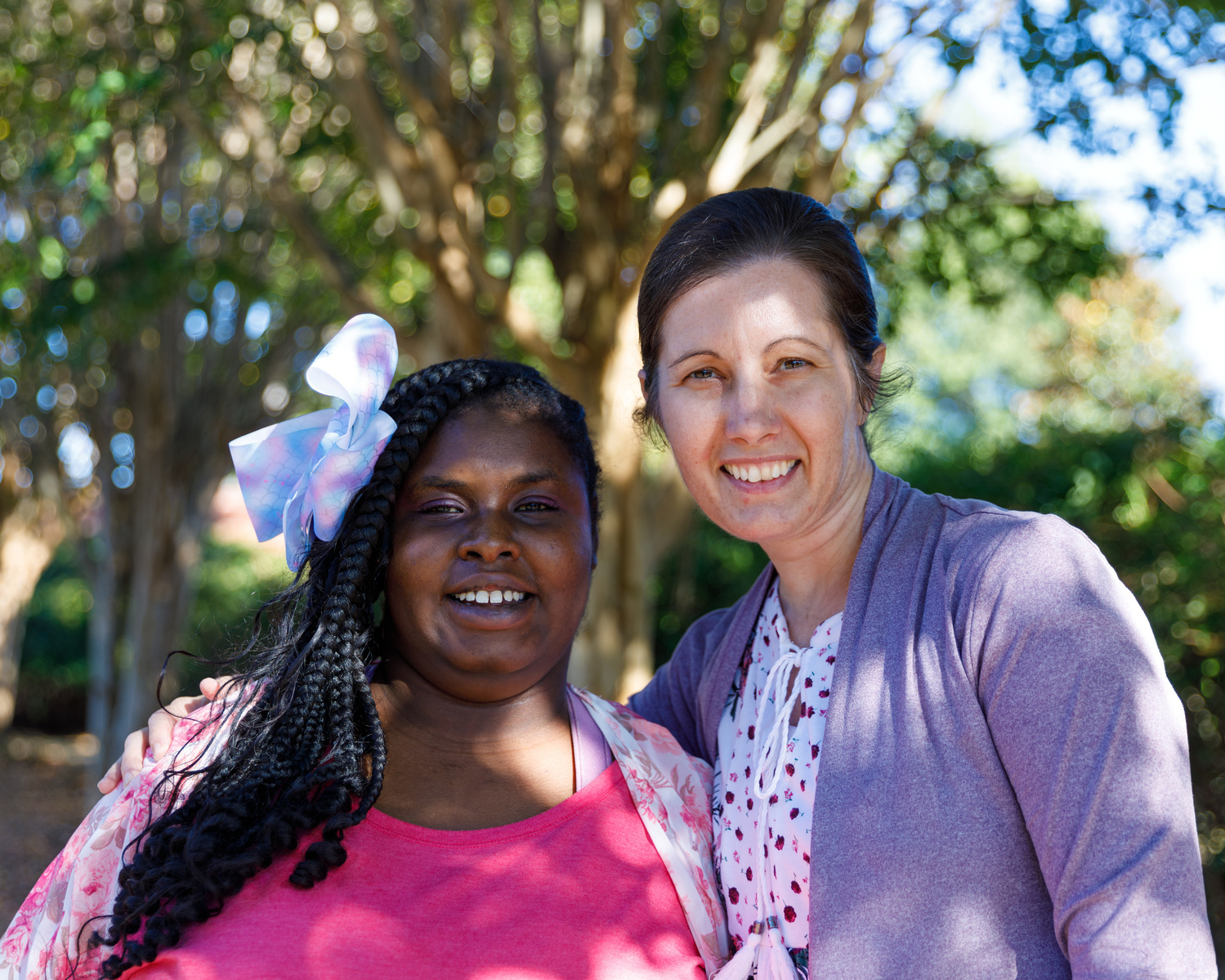 Mother and daughter stand outside under a tree on a sunny day. 