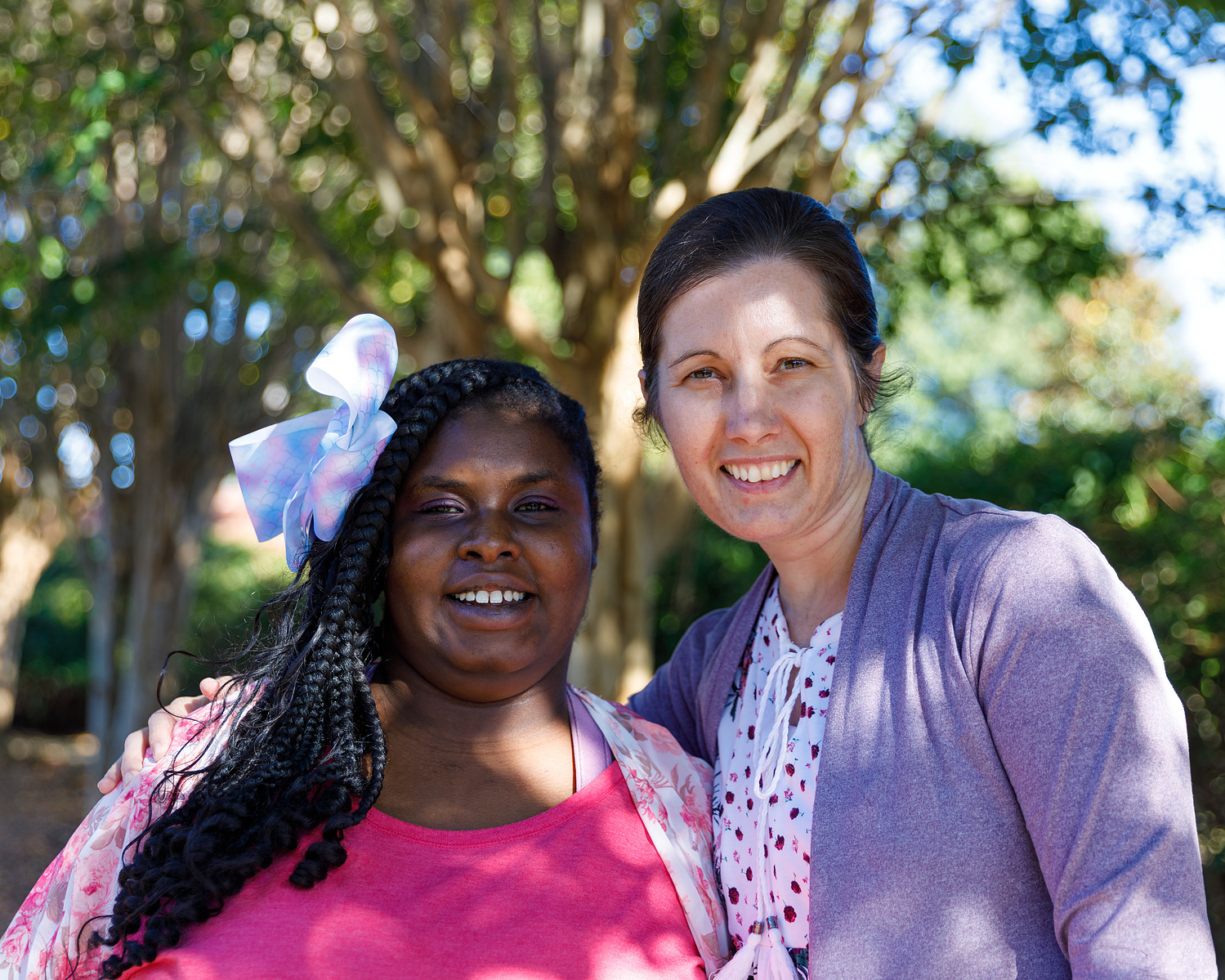 Mother and daughter stand outside under a tree on a sunny day. 