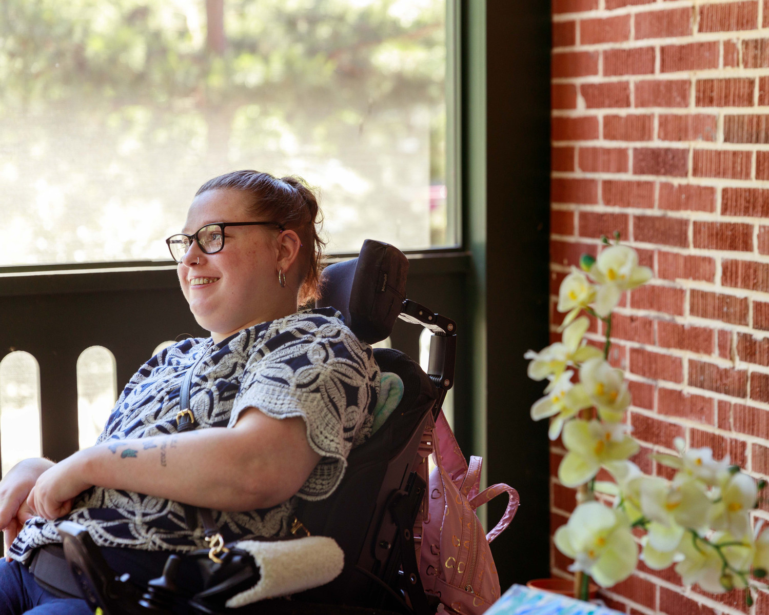 A young brunette woman in a patterned shirt and glasses sits on a screened porch in her wheelchair. 