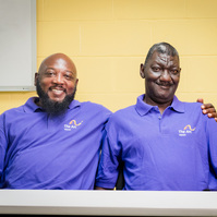 Two African American men wear matching purple polo shirts and sit at a table. 