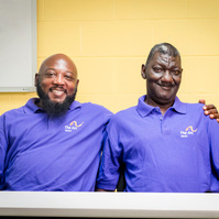 Two African American men wear matching purple polo shirts and sit at a table. 