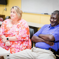 A man and a woman wear bright shirts and sit at a table in front of a yellow wall. 