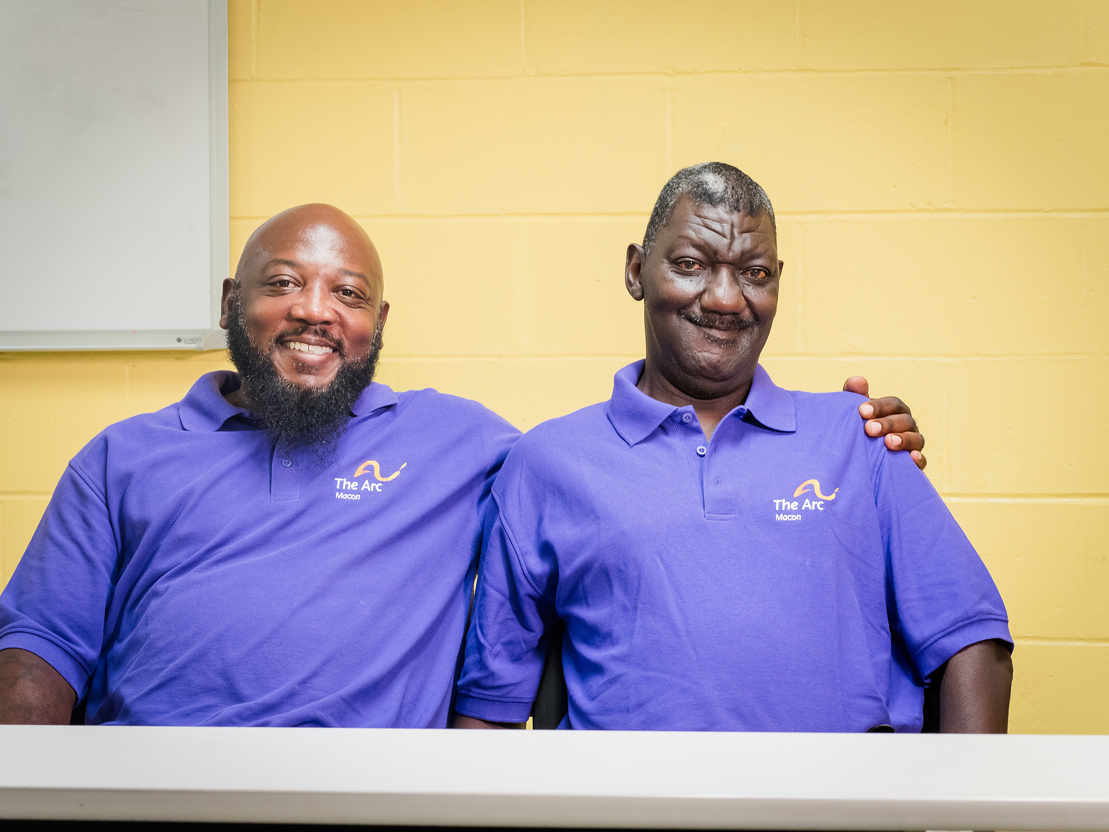 Two African American men wear matching purple polo shirts and sit at a table. 