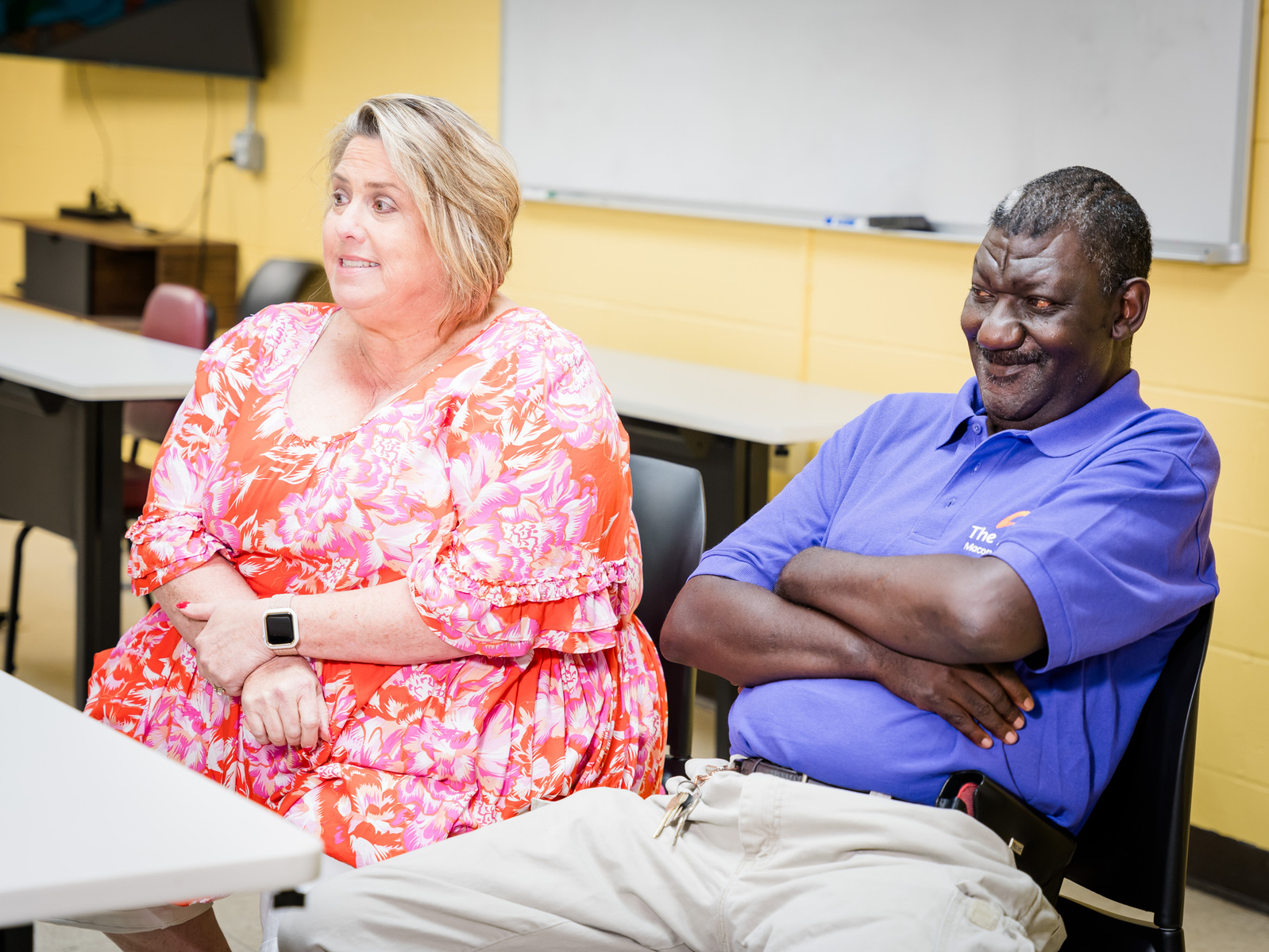 A man and a woman wear bright shirts and sit at a table in front of a yellow wall. 
