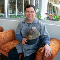 A young man in a grey collared shirt holds a baseball cap and sits on a sofa. 