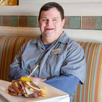 A young man in a grey shirt sits in a dining room at a table. 