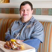 A young man in a grey shirt sits in a dining room at a table. 