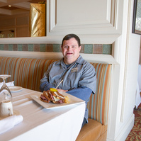 A young man in a grey shirt sits in a dining room at a table. 