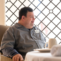 A young man in a grey shirt is shown sitting at a dining room table with a black leaded glass window behind him. 