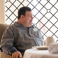 A young man in a grey shirt is shown sitting at a dining room table with a black leaded glass window behind him. 