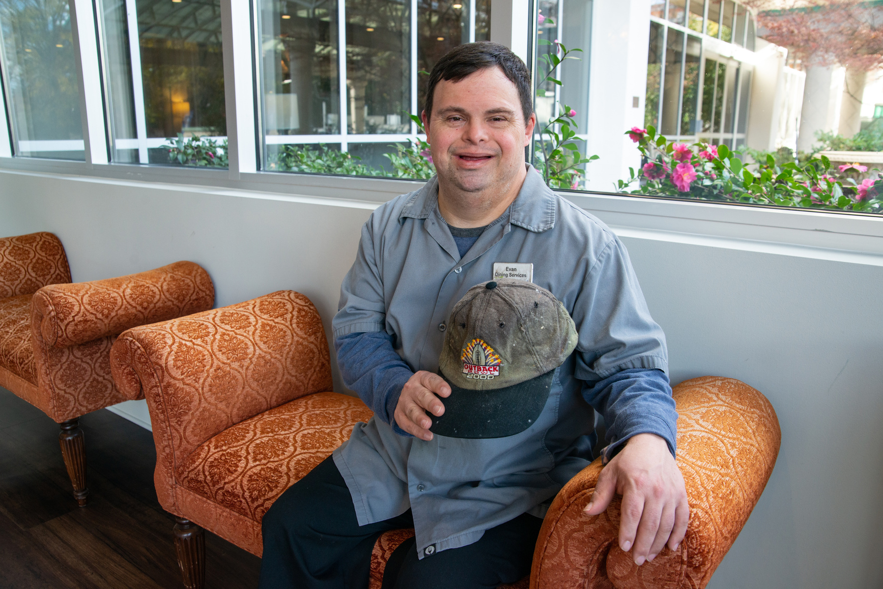 A young man in a grey collared shirt holds a baseball cap and sits on a sofa. 