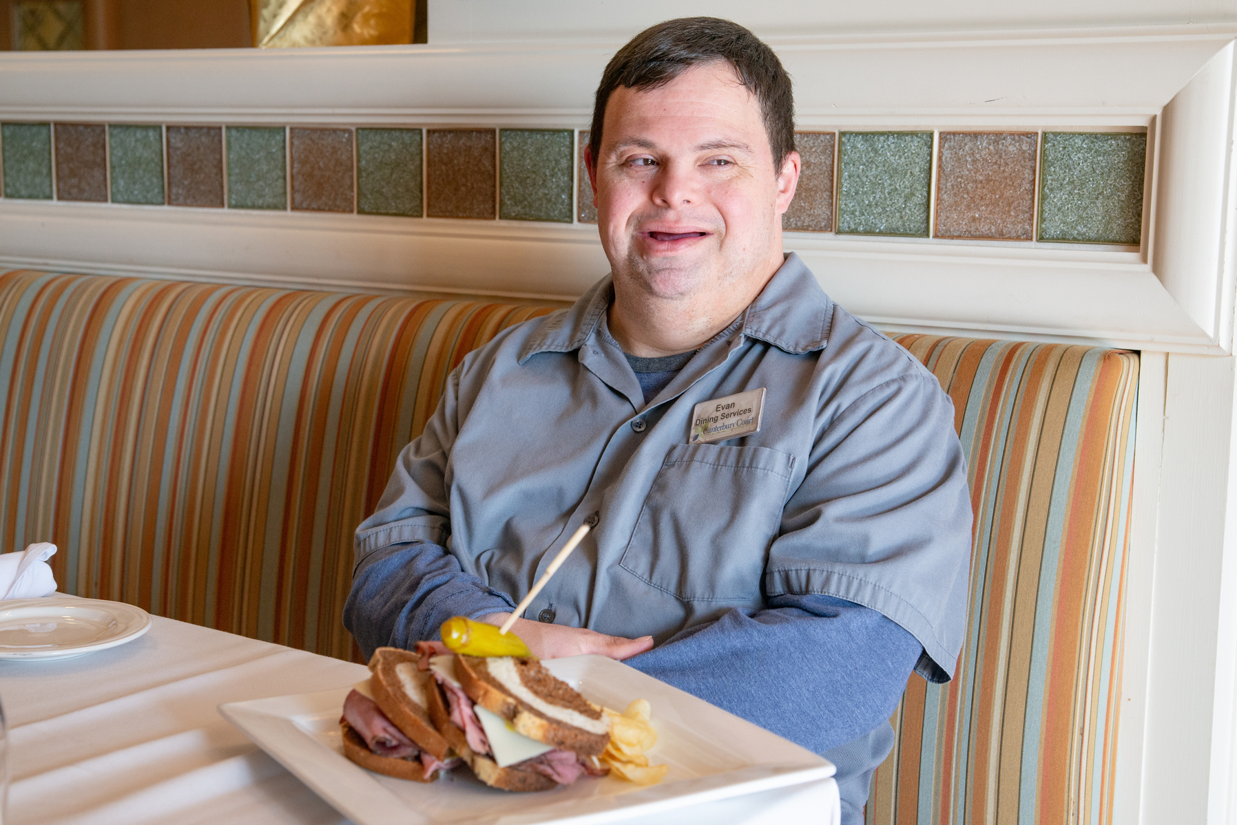 A young man in a grey shirt sits in a dining room at a table. 
