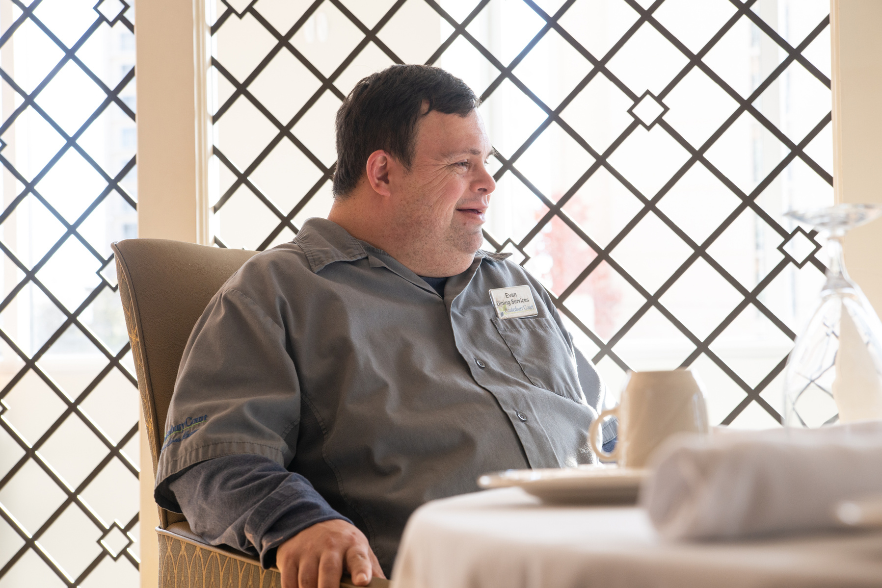 A young man in a grey shirt is shown sitting at a dining room table with a black leaded glass window behind him. 
