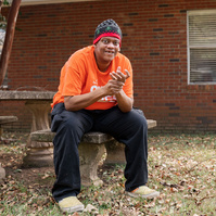 A man in an orange shirt and knit cap sits outside on a bench smiling.