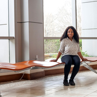 A young African American woman named Skye wears dark pants and a white blouse as she sits on a bench in front of a set of floor to ceiling windows. 