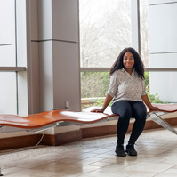 A young African American woman named Skye wears dark pants and a white blouse as she sits on a bench in front of a set of floor to ceiling windows. 