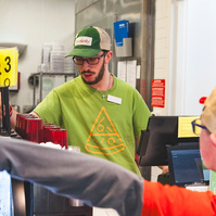 A bearded young man wears a green t-shirt and baseball cap while working behind a restaurant counter. 