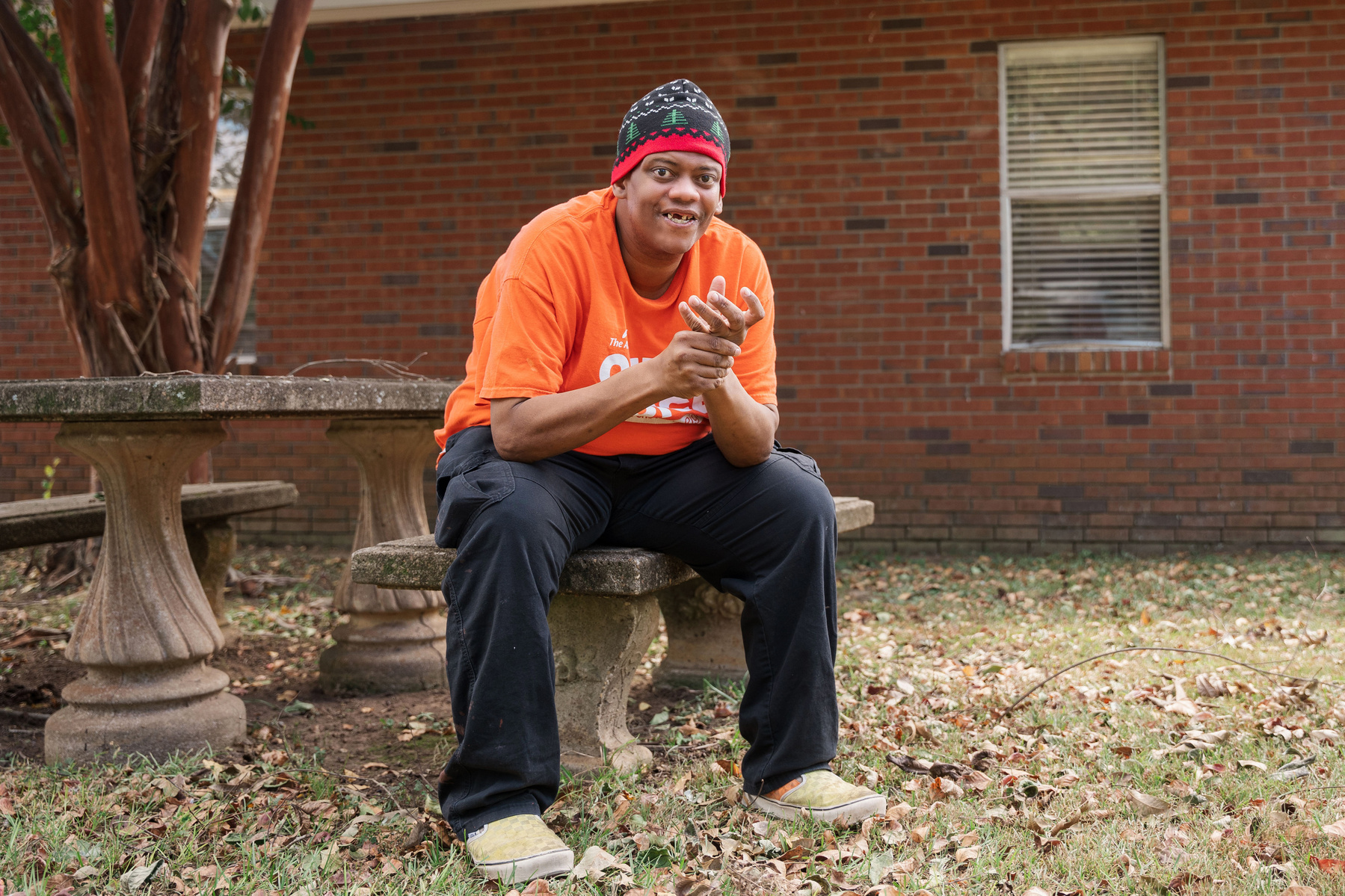 A man in an orange shirt and knit cap sits outside on a bench smiling.