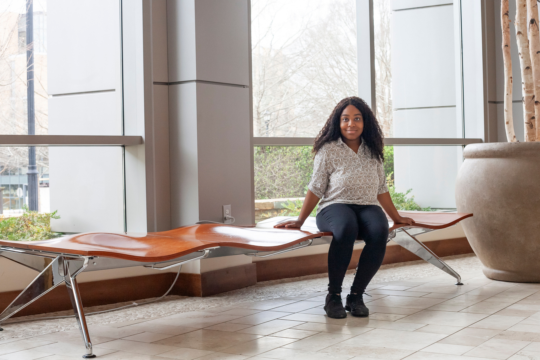 A young African American woman named Skye wears dark pants and a white blouse as she sits on a bench in front of a set of floor to ceiling windows. 
