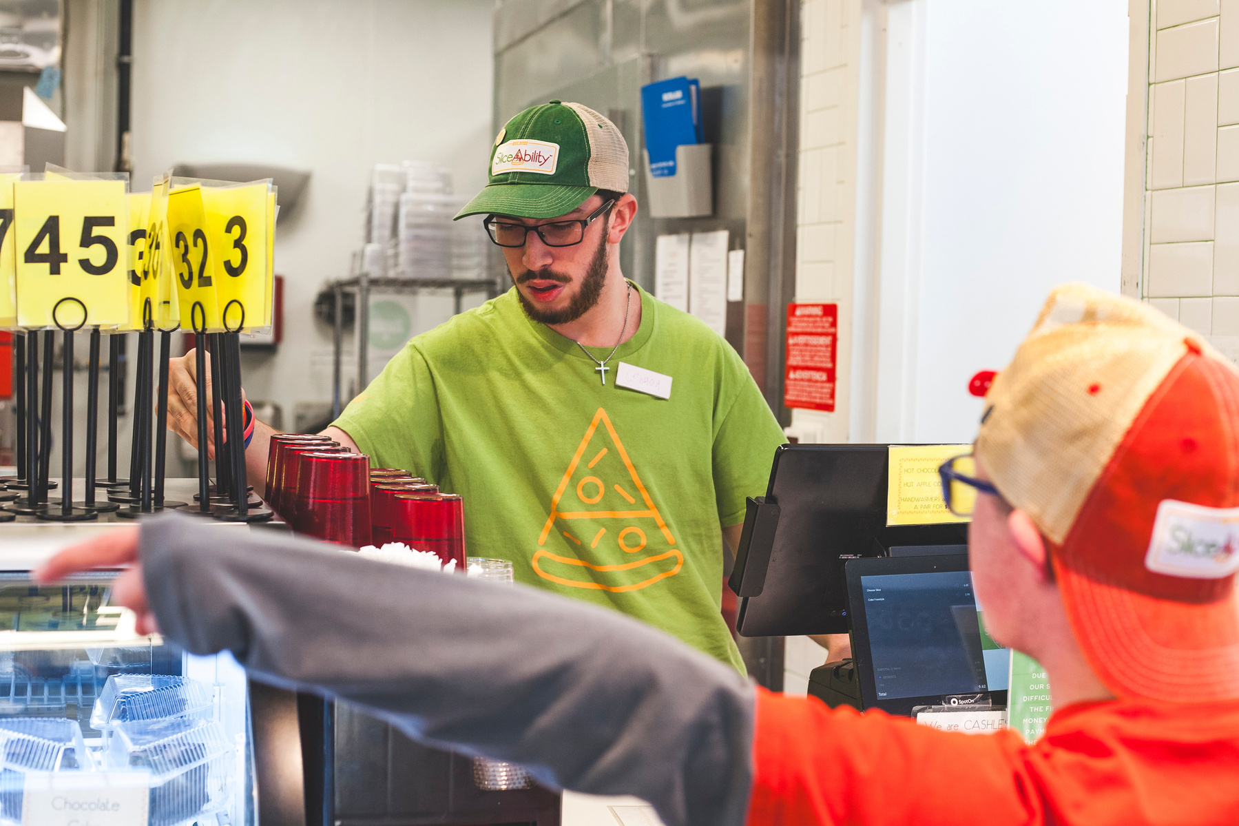 A bearded young man wears a green t-shirt and baseball cap while working behind a restaurant counter. 