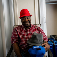 African American man in a plaid shirt and red hat smiles in his living room while holding a stack of hats.
