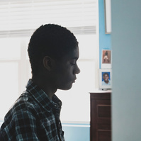 A young African American man in a plaid shirt sits in front of a window in his room. 