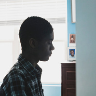 A young African American man in a plaid shirt sits in front of a window in his room. 