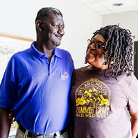 An African American man wearing a purple polo shirt hugs an African American woman with braid and glasses. 