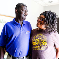 An African American man wearing a purple polo shirt hugs an African American woman with braid and glasses. 