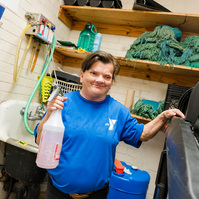A woman with brown hair wears a blue t-shirt and holds a spray bottle. 