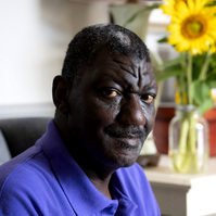 A middle aged African American man wears a purple polo shirt and sits in front of a vase full of sunflowers. 