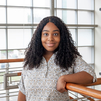 A young African American woman wearing a white blouse leans against a railing in front of a large window. 