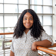 A young African American woman wearing a white blouse leans against a railing in front of a large window. 