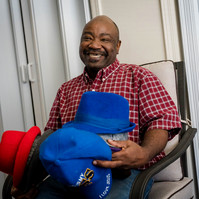 A man sits in a plaid shirt and holds a stack of hats. 