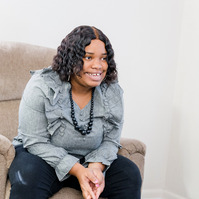 A young woman with dark hair wears a grey blouse and a black necklace. She is sitting on a recliner in the living room. 