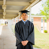 A young African American male wears a graduation cap and gown while standing in a walkway. 