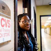 Young African American woman wears braids and stands next to the CVS Retail Training Classroom. 