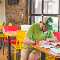 A bearded young man wears a green hat and t-shirt works at a restaurant table. 