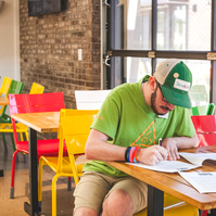 A bearded young man wears a green hat and t-shirt works at a restaurant table. 