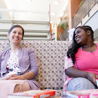 Mother and daughter sit on a patterned sofa and smile at each other. 
